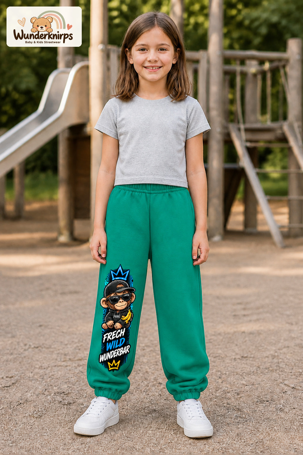 Mädchen trägt grüne Jogginghose mit Frech wild Affe-Print, von vorn auf dem Spielplatz.