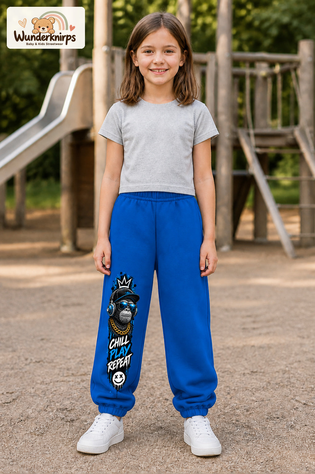 Mädchen trägt blaue Jogginghose mit Chill Play Affe-Print, von vorn auf dem Spielplatz.