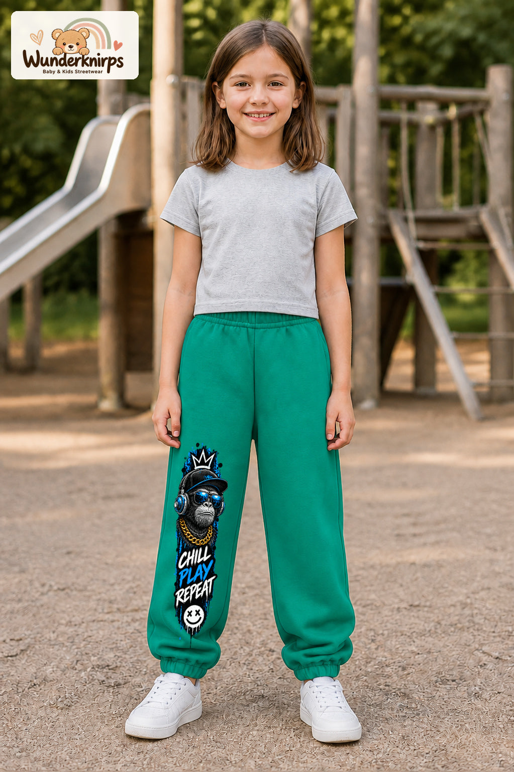 Mädchen trägt grüne Jogginghose mit Chill Play Affe-Print, von vorn auf dem Spielplatz.