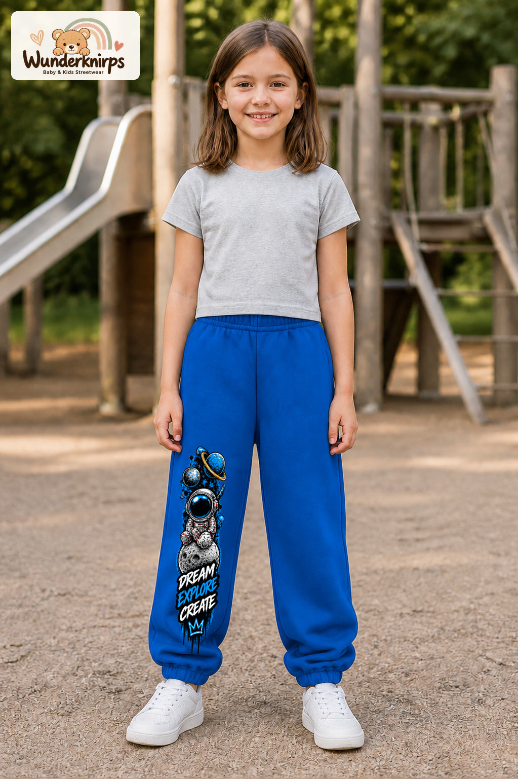 Mädchen trägt blaue Jogginghose mit Dream Explore Create-Print, von vorn auf dem Spielplatz.