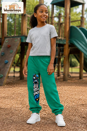 Mädchen trägt grüne Jogginghose mit Space Kid Mondreise-Print, leicht seitlich auf dem Spielplatz.
