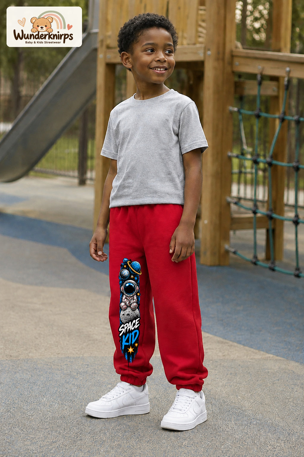 Junge trägt rote Jogginghose mit Space Kid Mondreise-Print, leicht seitlich auf dem Spielplatz.