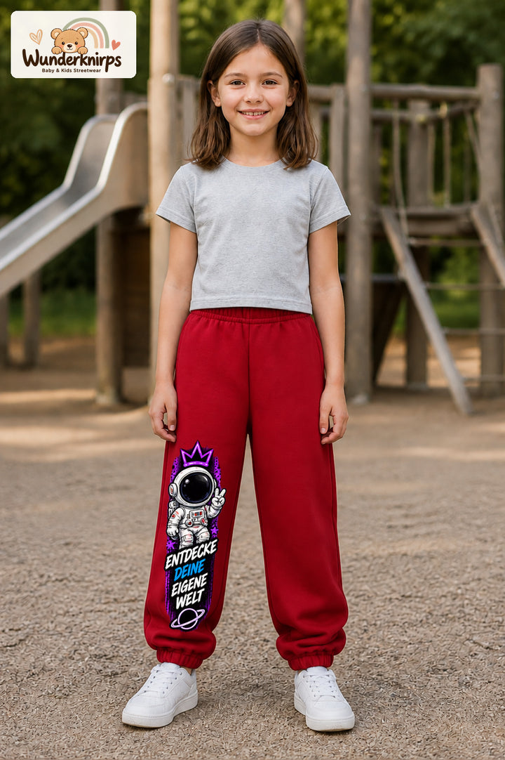 Mädchen trägt rote Jogginghose mit Eigene Welt Astronaut-Print, von vorn auf dem Spielplatz.