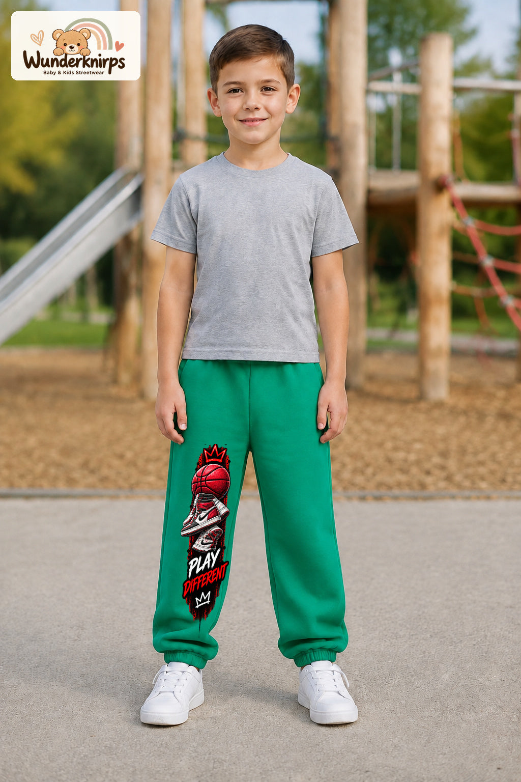 Junge trägt grüne Jogginghose mit Play Different Basketball-Print, von vorn auf dem Spielplatz.