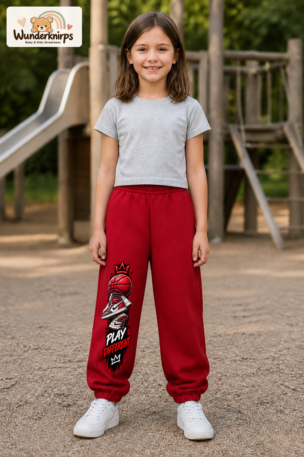 Mädchen trägt rote Jogginghose mit Play Different Basketball-Print, von vorn auf dem Spielplatz.