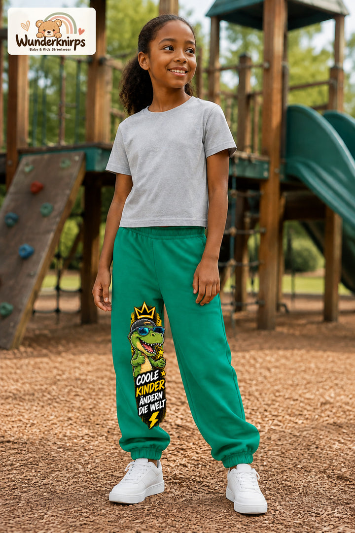 Mädchen trägt grüne Jogginghose mit Coole Kinder Sonnenbrillen-Dino-Print, leicht seitlich auf dem Spielplatz.