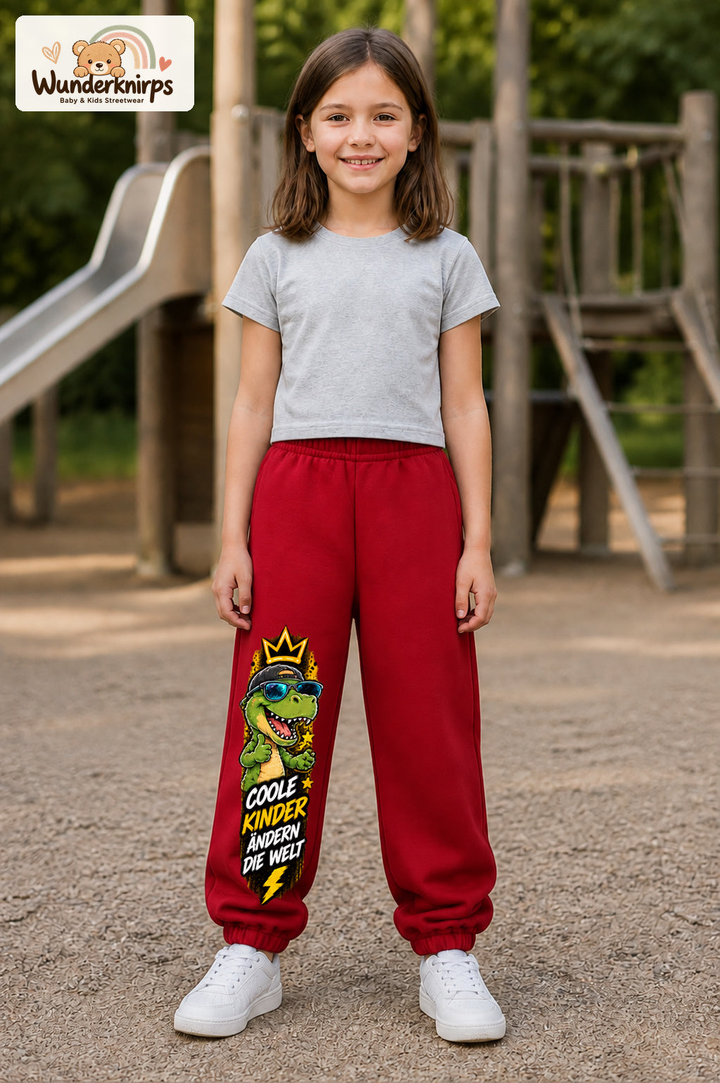 Mädchen trägt rote Jogginghose mit Coole Kinder Sonnenbrillen-Dino-Print, von vorn auf dem Spielplatz.