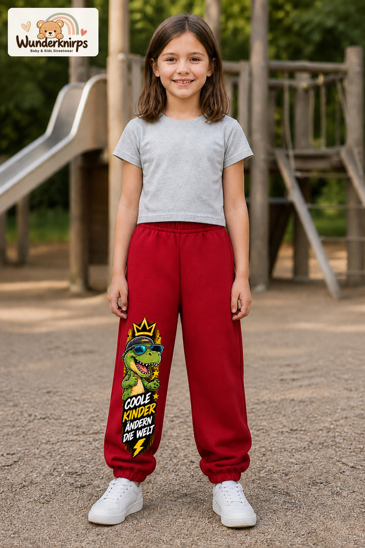 Mädchen trägt rote Jogginghose mit Coole Kinder Sonnenbrillen-Dino-Print, von vorn auf dem Spielplatz.