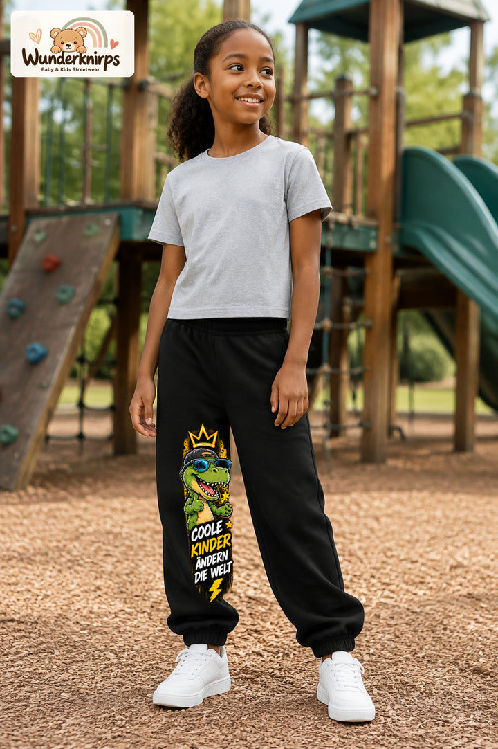 Mädchen trägt schwarze Jogginghose mit Coole Kinder Sonnenbrillen-Dino-Print, leicht seitlich auf dem Spielplatz.