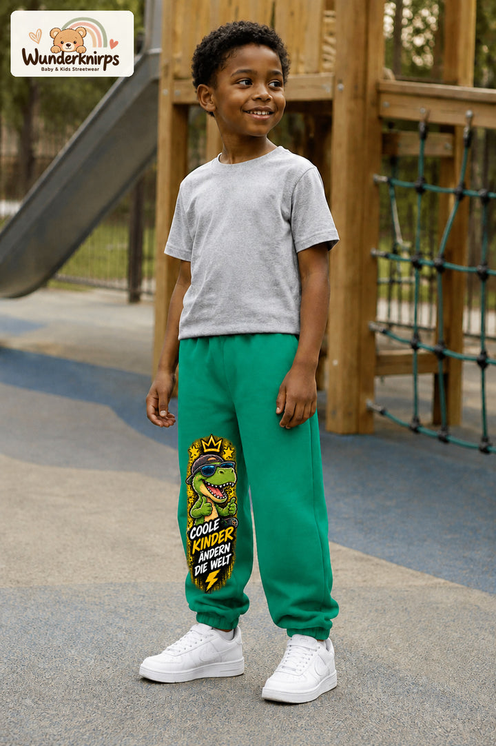 Junge trägt grüne Jogginghose mit Coole Kinder Daumen-Dino-Print, leicht seitlich auf dem Spielplatz.