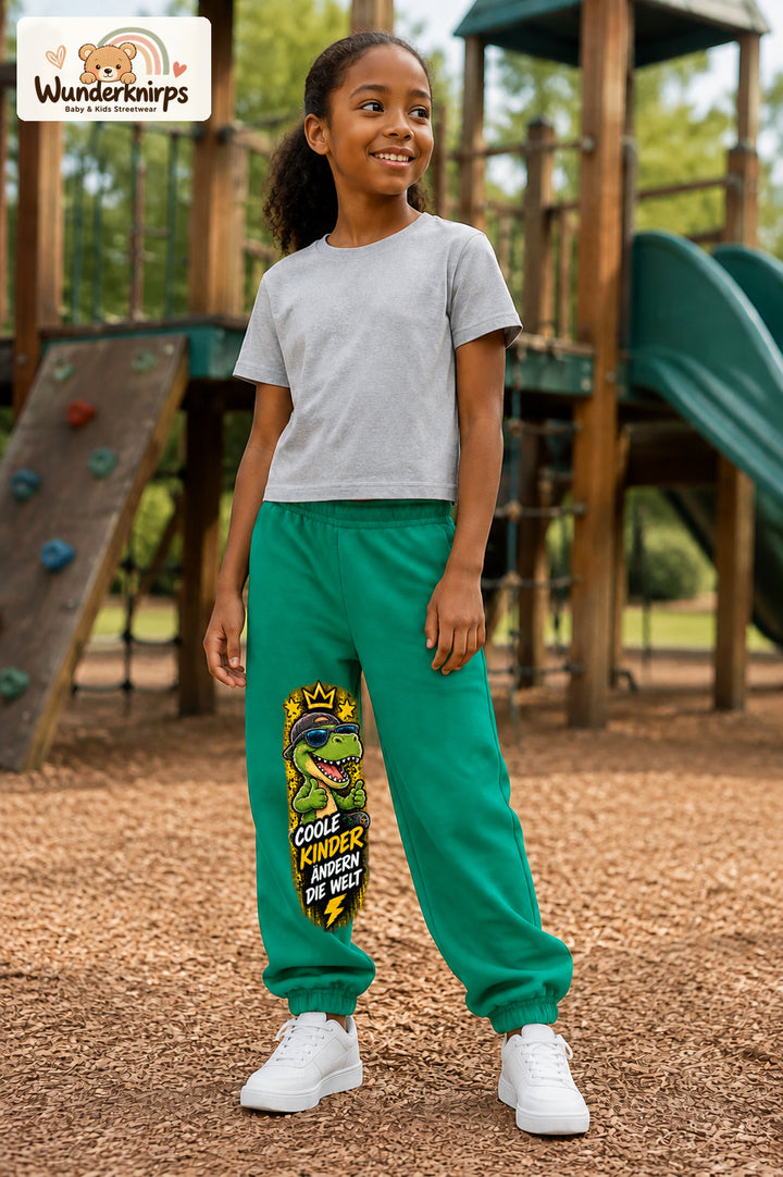Mädchen trägt grüne Jogginghose mit Coole Kinder Daumen-Dino-Print, leicht seitlich auf dem Spielplatz.