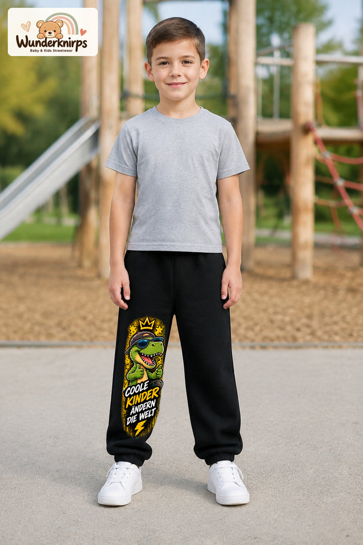 Junge trägt schwarze Jogginghose mit Coole Kinder Daumen-Dino-Print, von vorn auf dem Spielplatz.