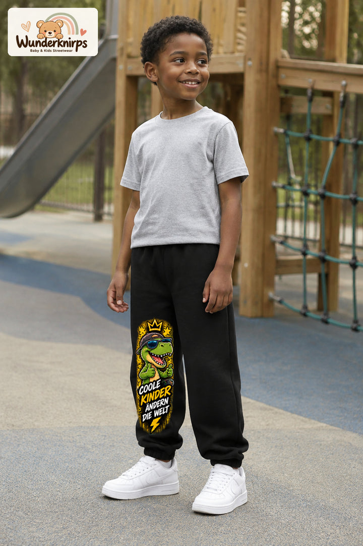Junge trägt schwarze Jogginghose mit Coole Kinder Daumen-Dino-Print, leicht seitlich auf dem Spielplatz.
