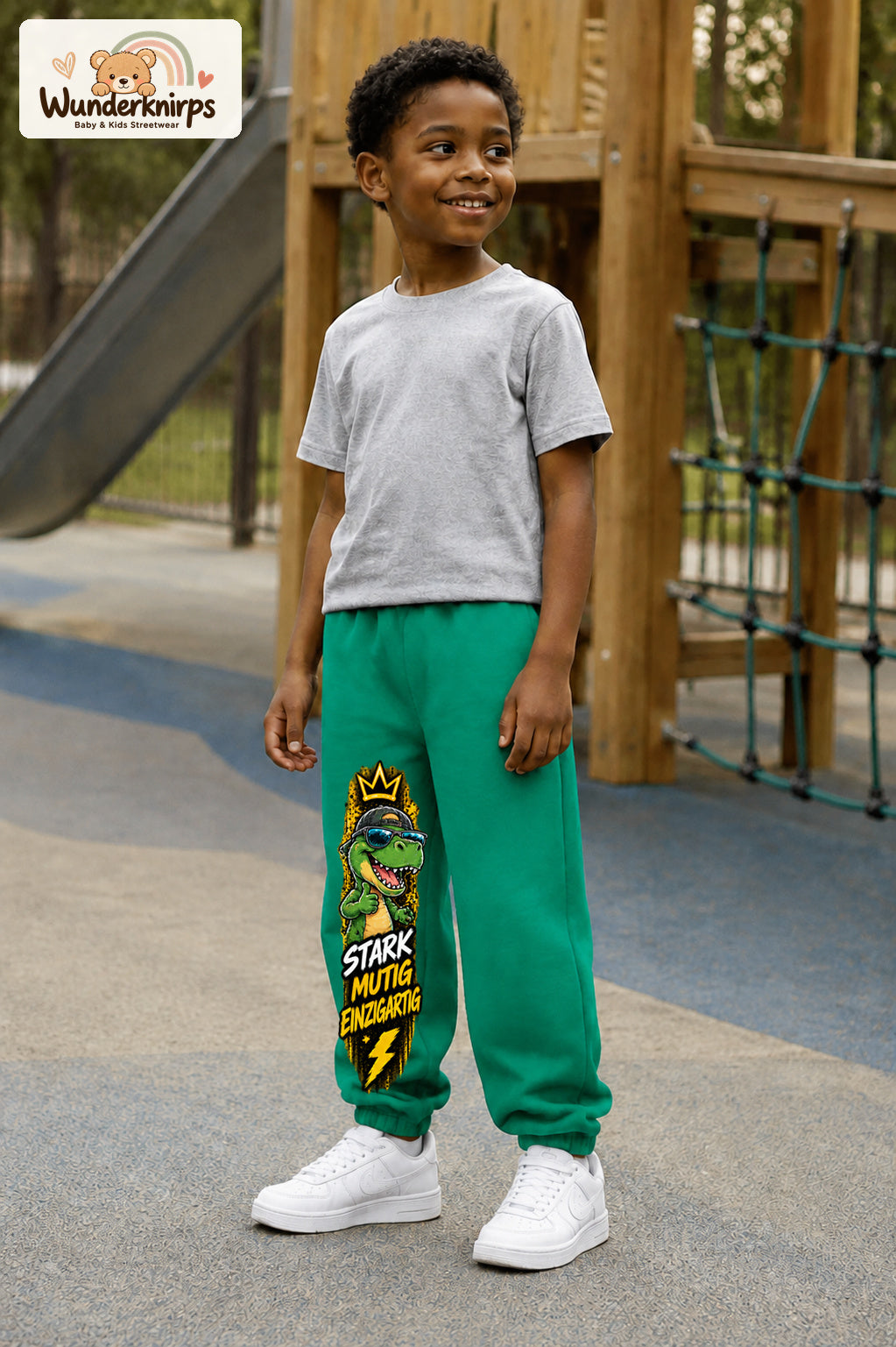 Junge trägt grüne Jogginghose mit Stark mutig Dino-Print, leicht seitlich auf dem Spielplatz.