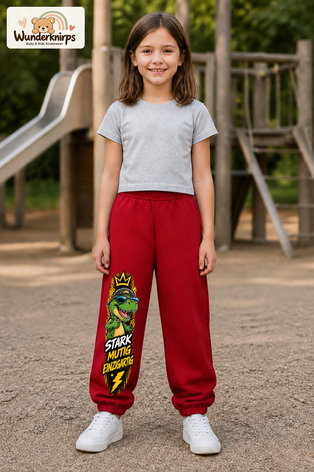 Mädchen trägt rote Jogginghose mit Stark mutig Dino-Print, von vorn auf dem Spielplatz.