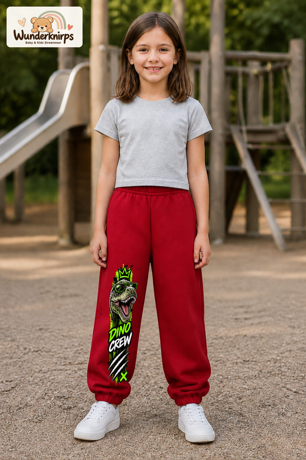 Mädchen trägt rote Jogginghose mit Dino Crew T-Rex-Print, von vorn auf dem Spielplatz.