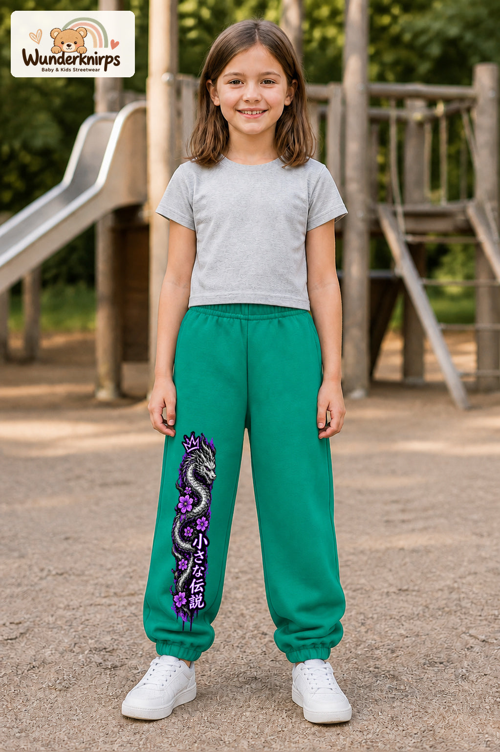 Mädchen trägt grüne Jogginghose mit Blütendrache Lila-Print, von vorn auf dem Spielplatz.