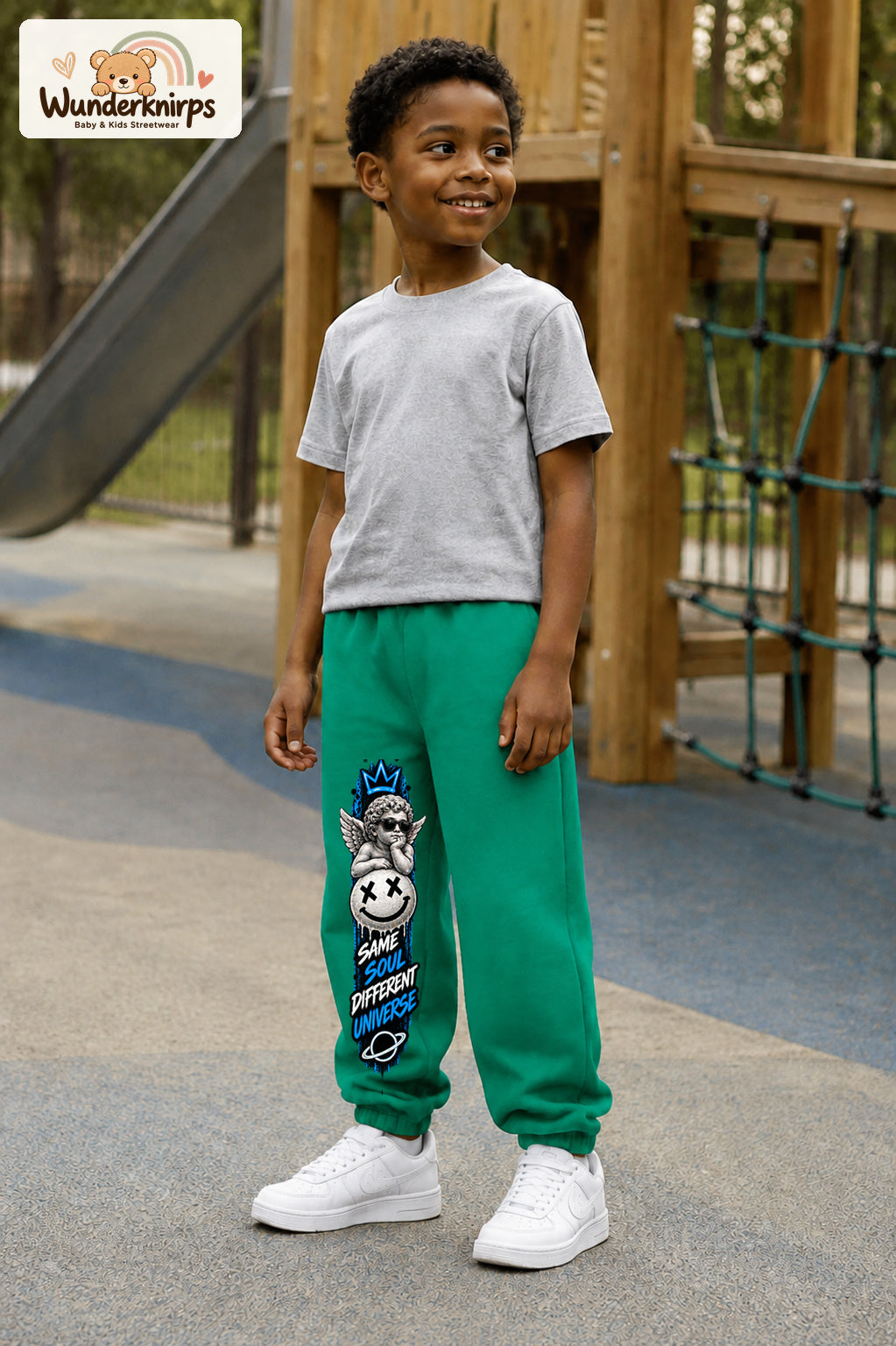 Junge trägt grüne Jogginghose mit Same Soul Engel-Print, leicht seitlich auf dem Spielplatz.