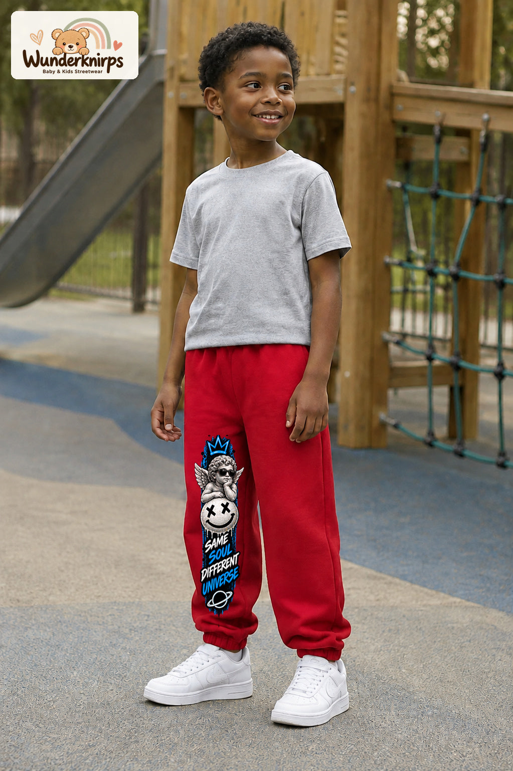 Junge trägt rote Jogginghose mit Same Soul Engel-Print, leicht seitlich auf dem Spielplatz.
