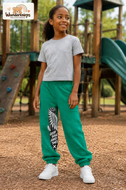 Mädchen trägt grüne Jogginghose mit Good Soul Flügel-Print, leicht seitlich auf dem Spielplatz.