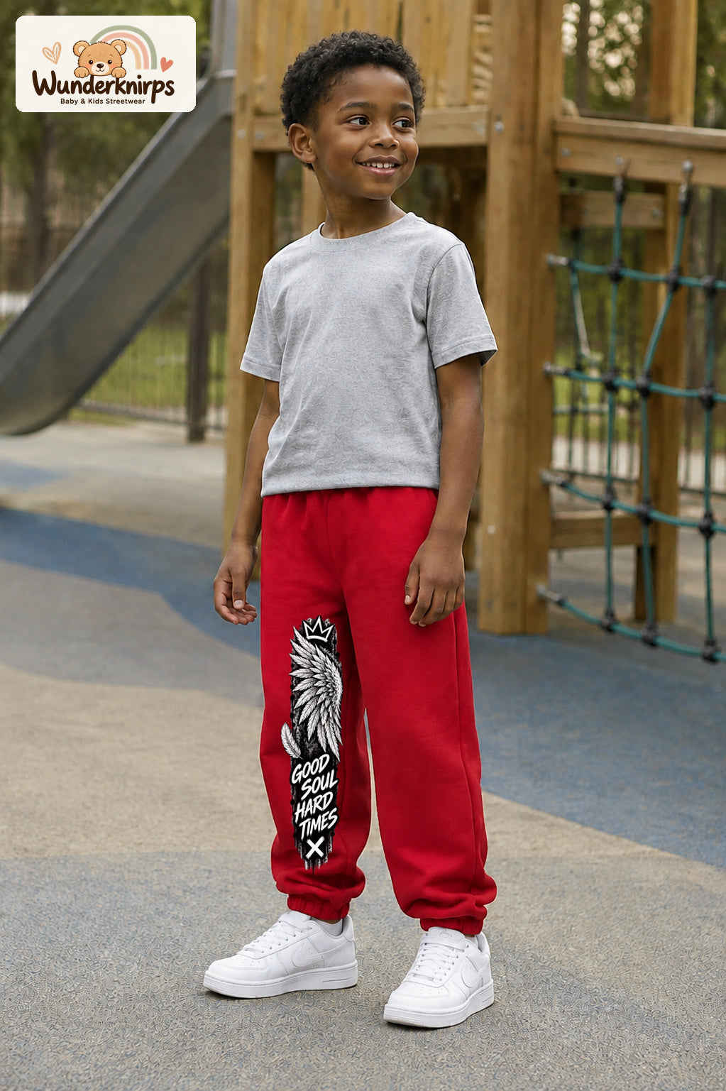 Junge trägt rote Jogginghose mit Good Soul Flügel-Print, leicht seitlich auf dem Spielplatz.
