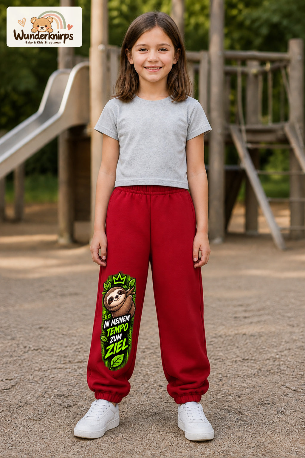 Mädchen trägt rote Jogginghose mit Mein Tempo Faultier-Print, von vorn auf dem Spielplatz.