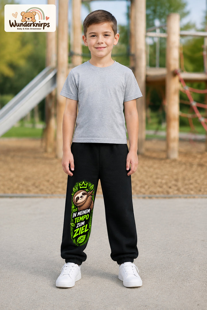 Junge trägt schwarze Jogginghose mit Mein Tempo Faultier-Print, von vorn auf dem Spielplatz.