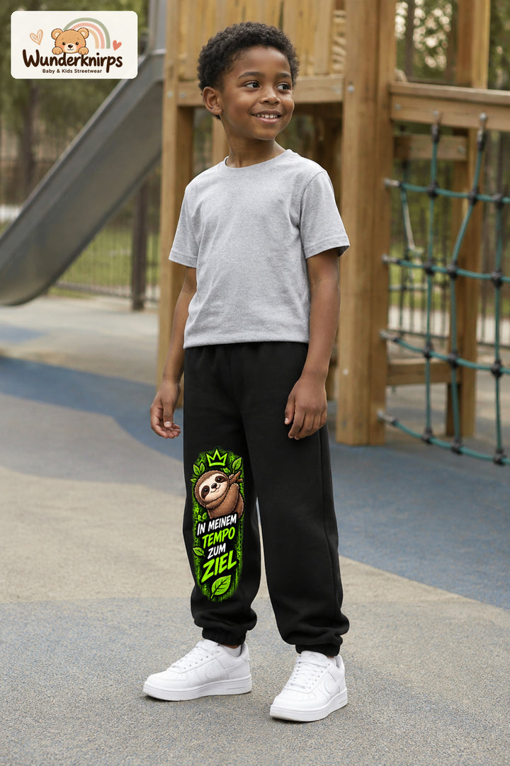 Junge trägt schwarze Jogginghose mit Mein Tempo Faultier-Print, leicht seitlich auf dem Spielplatz.