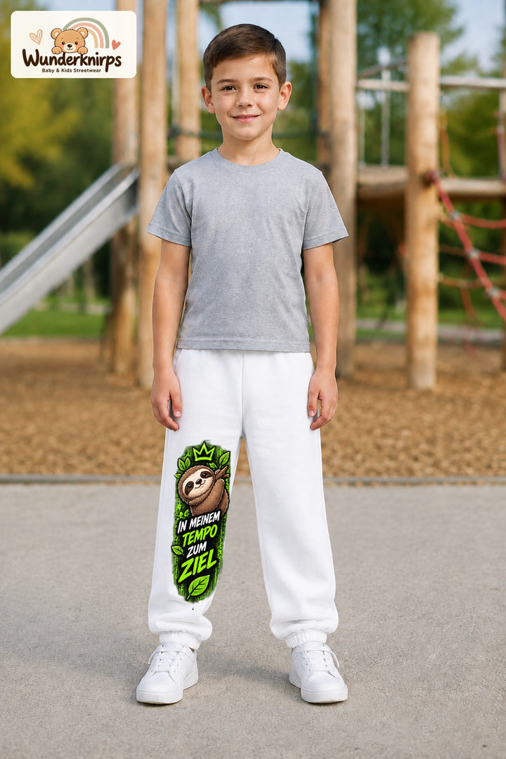 Junge trägt weiße Jogginghose mit Mein Tempo Faultier-Print, von vorn auf dem Spielplatz.