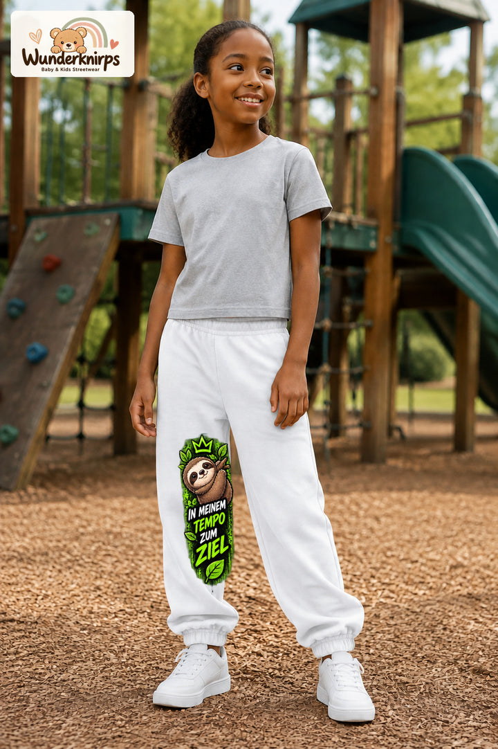 Mädchen trägt weiße Jogginghose mit Mein Tempo Faultier-Print, leicht seitlich auf dem Spielplatz.