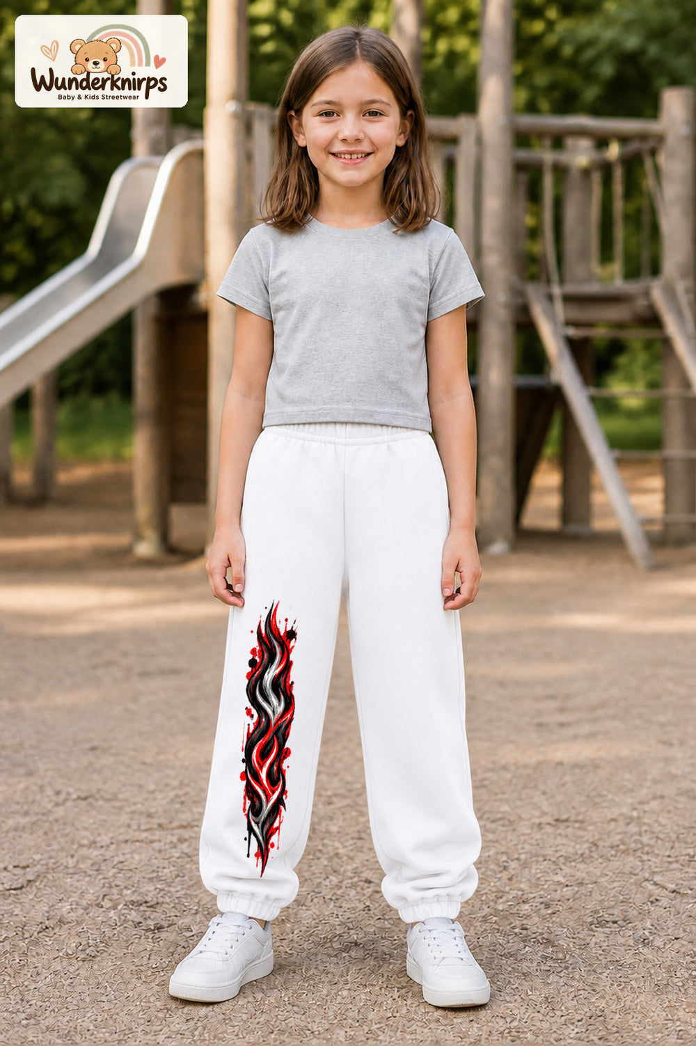 Mädchen trägt weiße Jogginghose mit Rote Flammen-Print, von vorn auf dem Spielplatz.