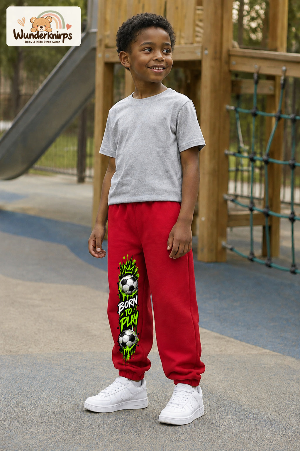 Junge trägt rote Jogginghose mit Born to Play Fußball-Print, leicht seitlich auf dem Spielplatz.