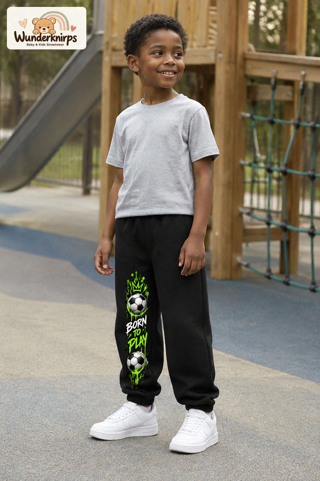 Junge trägt schwarze Jogginghose mit Born to Play Fußball-Print, leicht seitlich auf dem Spielplatz.
