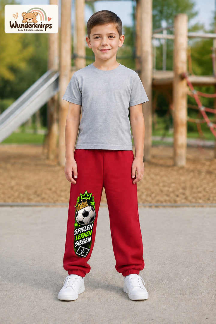 Junge trägt rote Jogginghose mit Spielen Lernen Fußball-Print, von vorn auf dem Spielplatz.