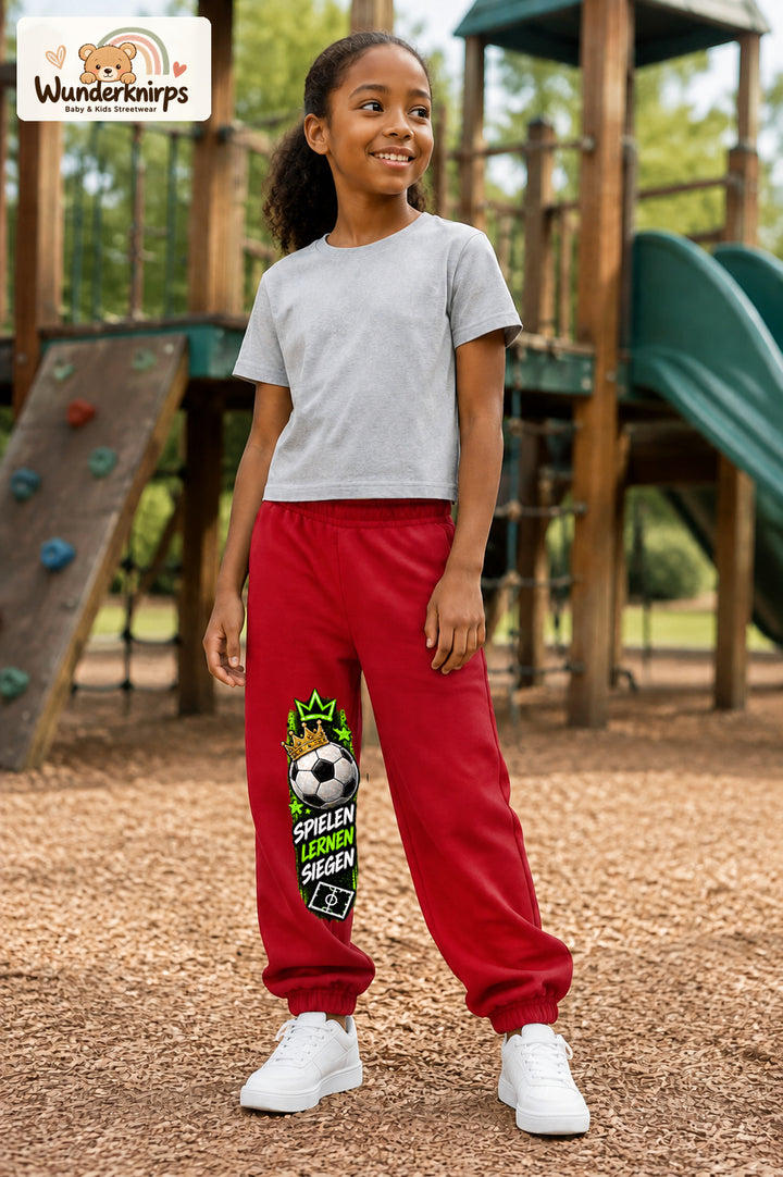 Mädchen trägt rote Jogginghose mit Spielen Lernen Fußball-Print, leicht seitlich auf dem Spielplatz.