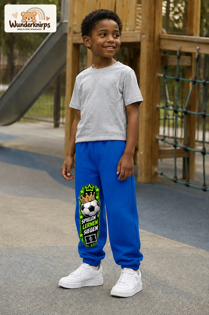 Junge trägt blaue Jogginghose mit Siegen Fußballstern-Print, leicht seitlich auf dem Spielplatz.