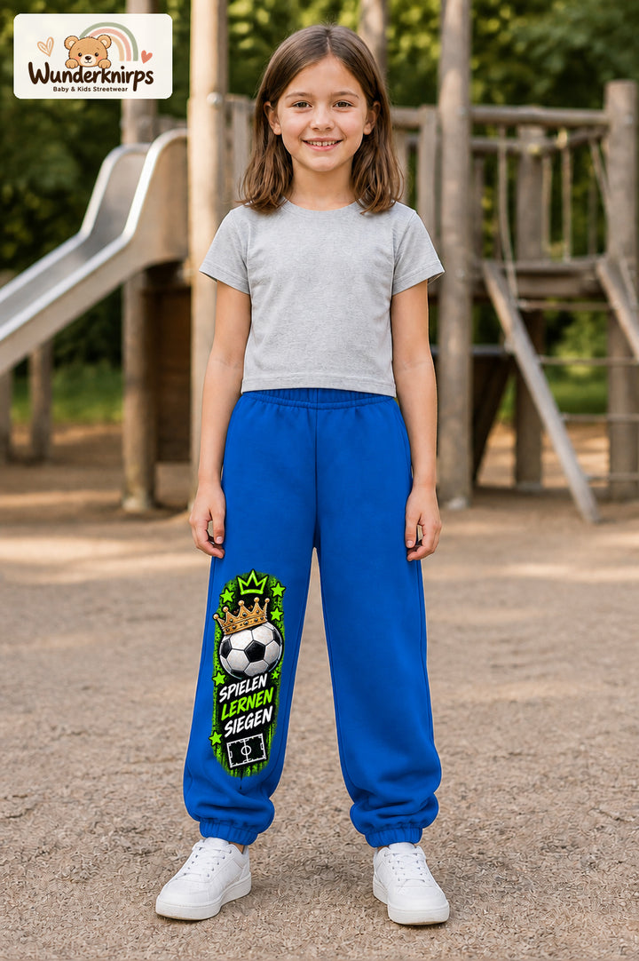Mädchen trägt blaue Jogginghose mit Siegen Fußballstern-Print, von vorn auf dem Spielplatz.