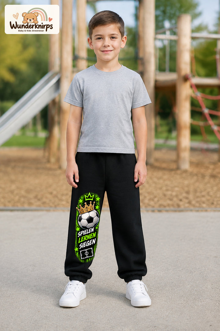 Junge trägt schwarze Jogginghose mit Siegen Fußballstern-Print, von vorn auf dem Spielplatz.