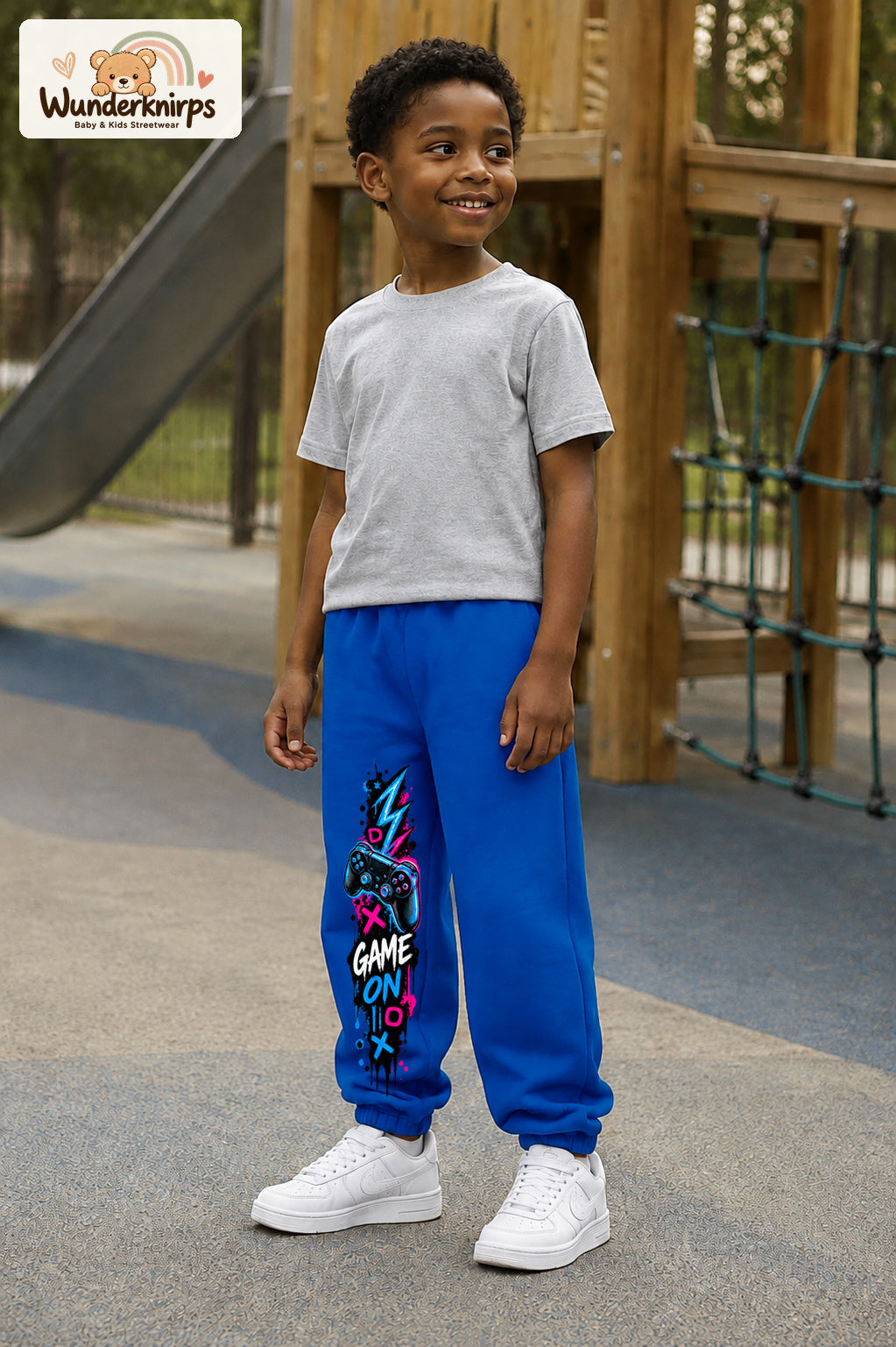 Junge trägt blaue Jogginghose mit Game On Controller-Print, leicht seitlich auf dem Spielplatz.