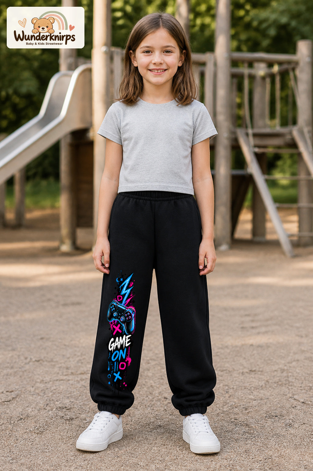 Mädchen trägt schwarze Jogginghose mit Game On Controller-Print, von vorn auf dem Spielplatz.