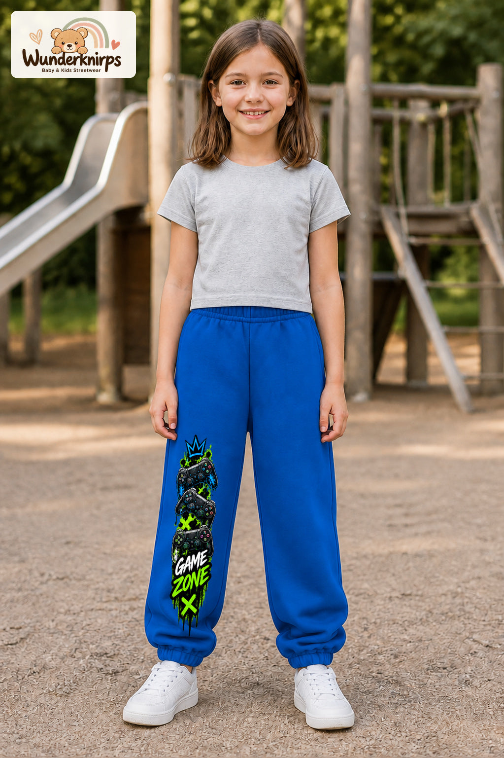 Mädchen trägt blaue Jogginghose mit Game Zone Controller-Print, von vorn auf dem Spielplatz.