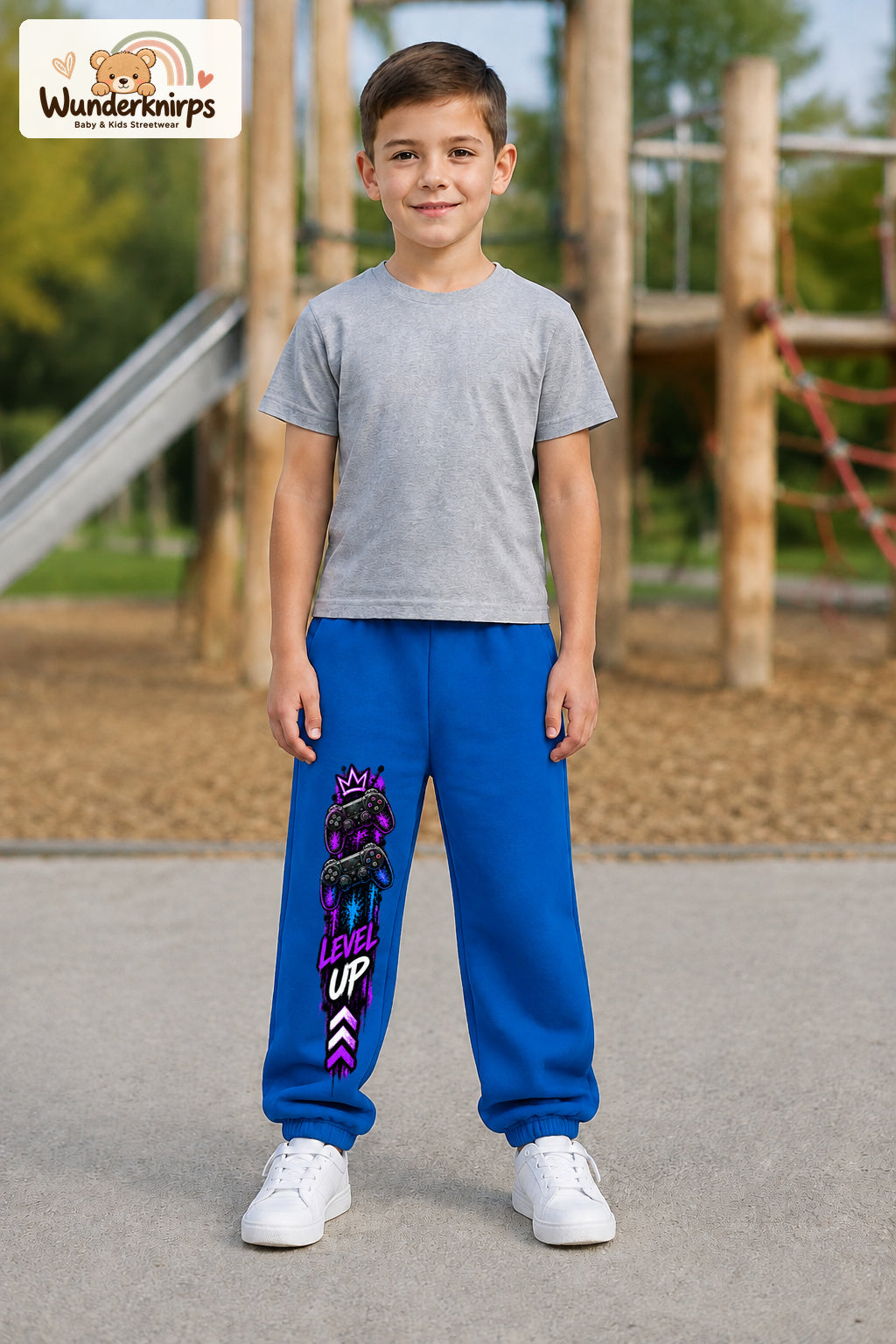 Junge trägt blaue Jogginghose mit Level Up Gaming-Print, von vorn auf dem Spielplatz.