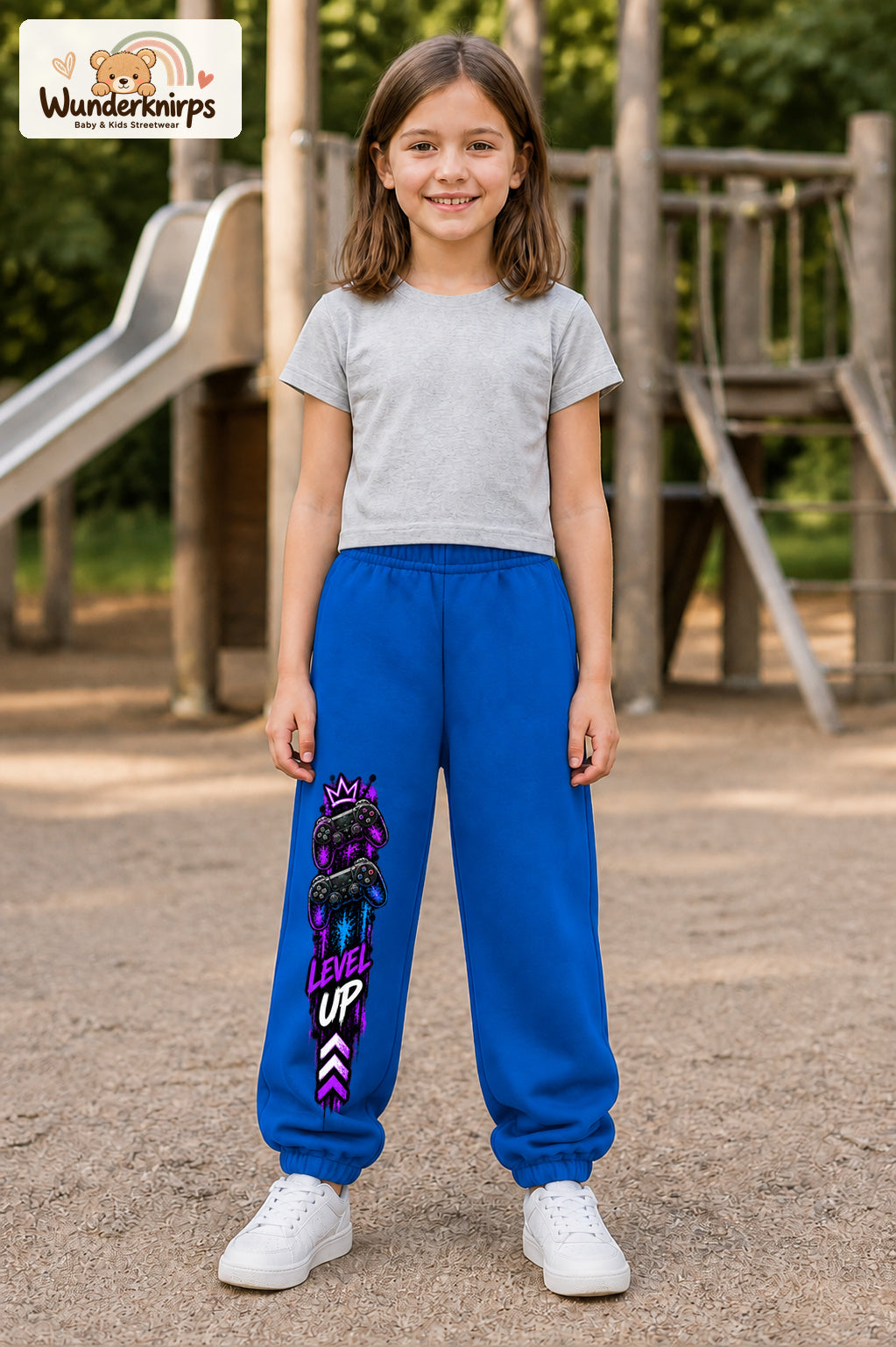 Mädchen trägt blaue Jogginghose mit Level Up Gaming-Print, von vorn auf dem Spielplatz.