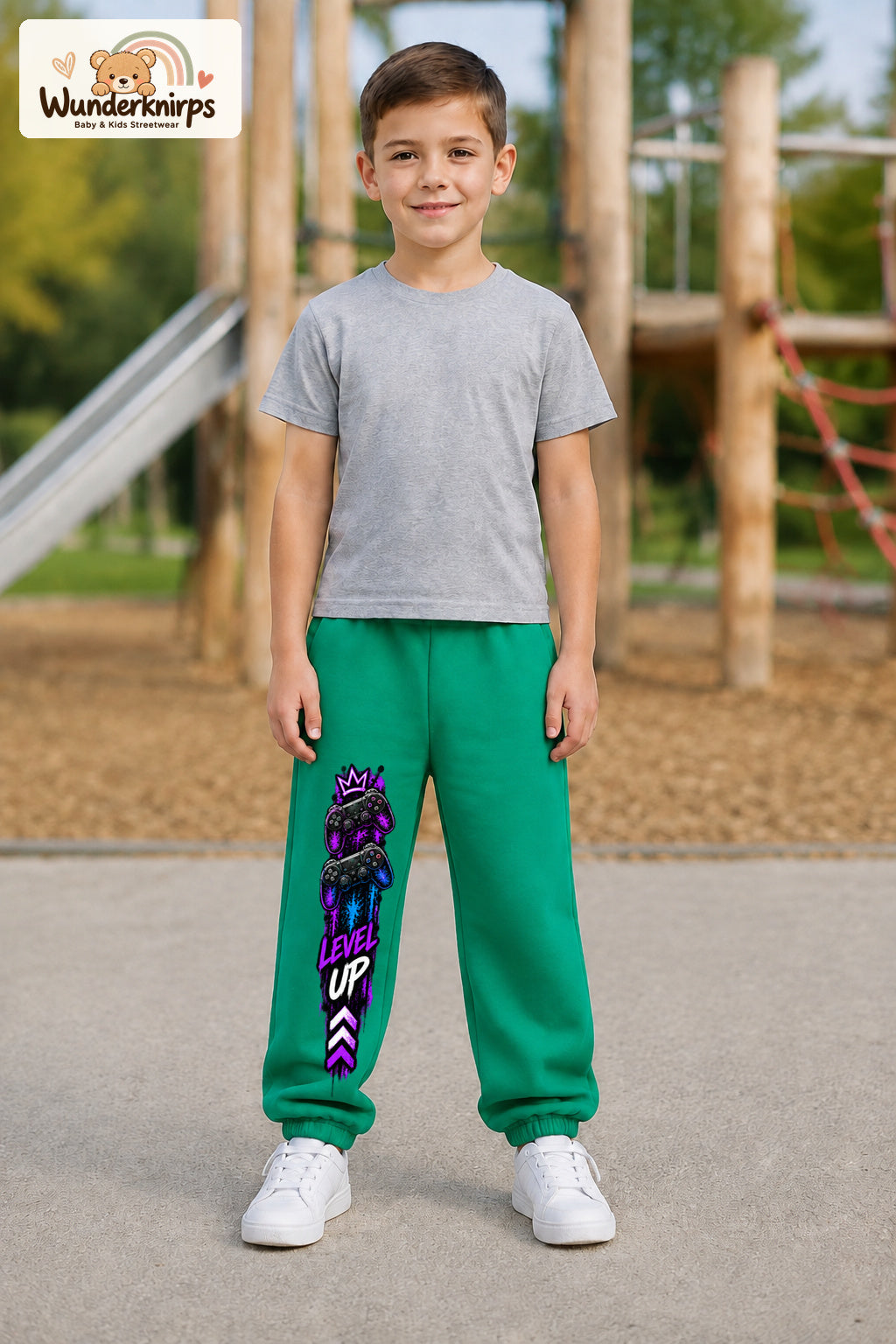 Junge trägt grüne Jogginghose mit Level Up Gaming-Print, von vorn auf dem Spielplatz.