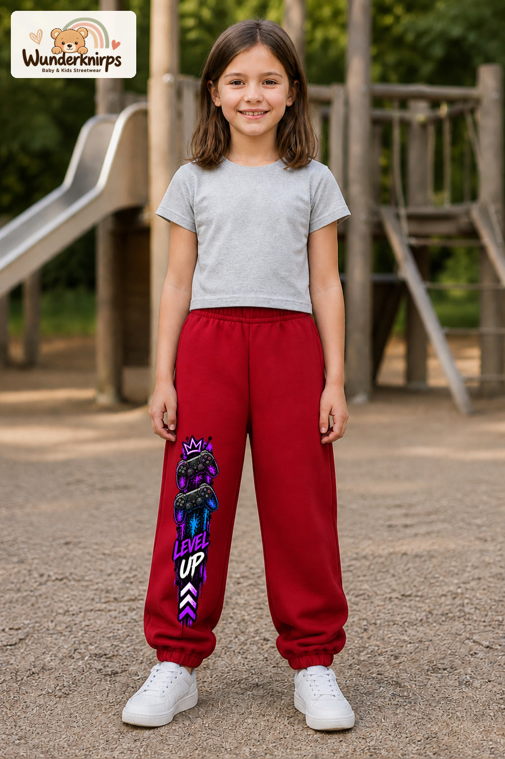 Mädchen trägt rote Jogginghose mit Level Up Gaming-Print, von vorn auf dem Spielplatz.
