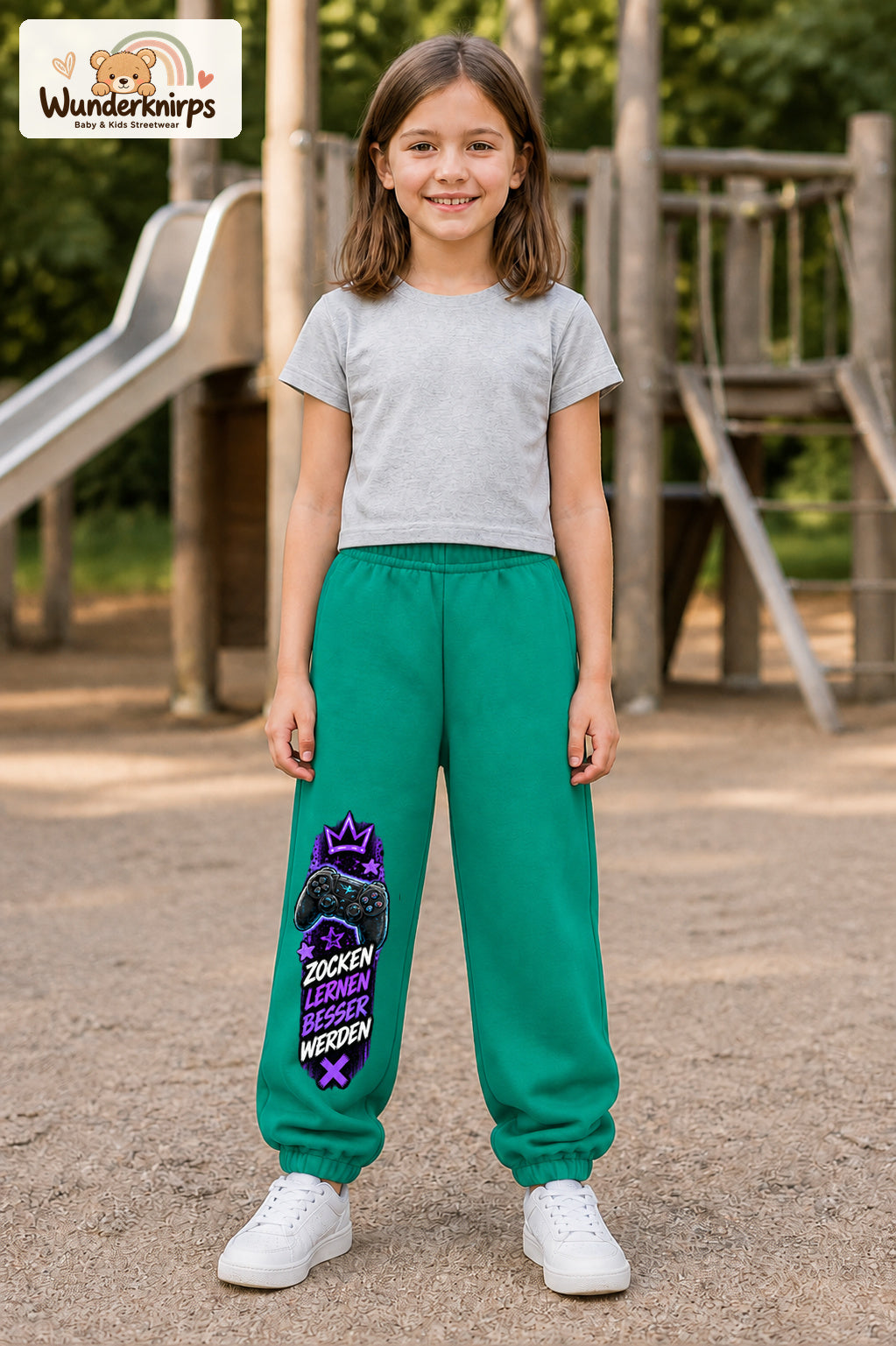 Mädchen trägt grüne Jogginghose mit Besser werden Gamepad-Print, von vorn auf dem Spielplatz.