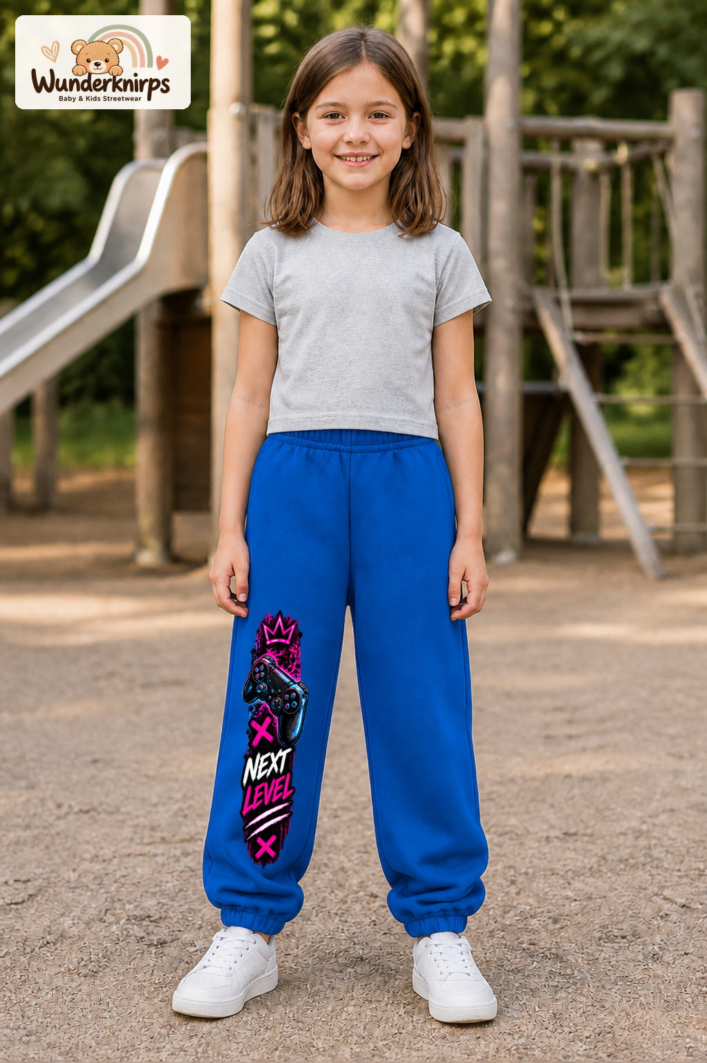 Mädchen trägt blaue Jogginghose mit Next Level Controller-Print, von vorn auf dem Spielplatz.