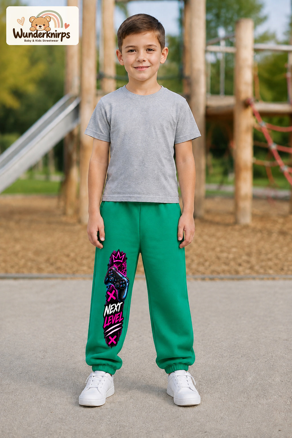 Junge trägt grüne Jogginghose mit Next Level Controller-Print, von vorn auf dem Spielplatz.