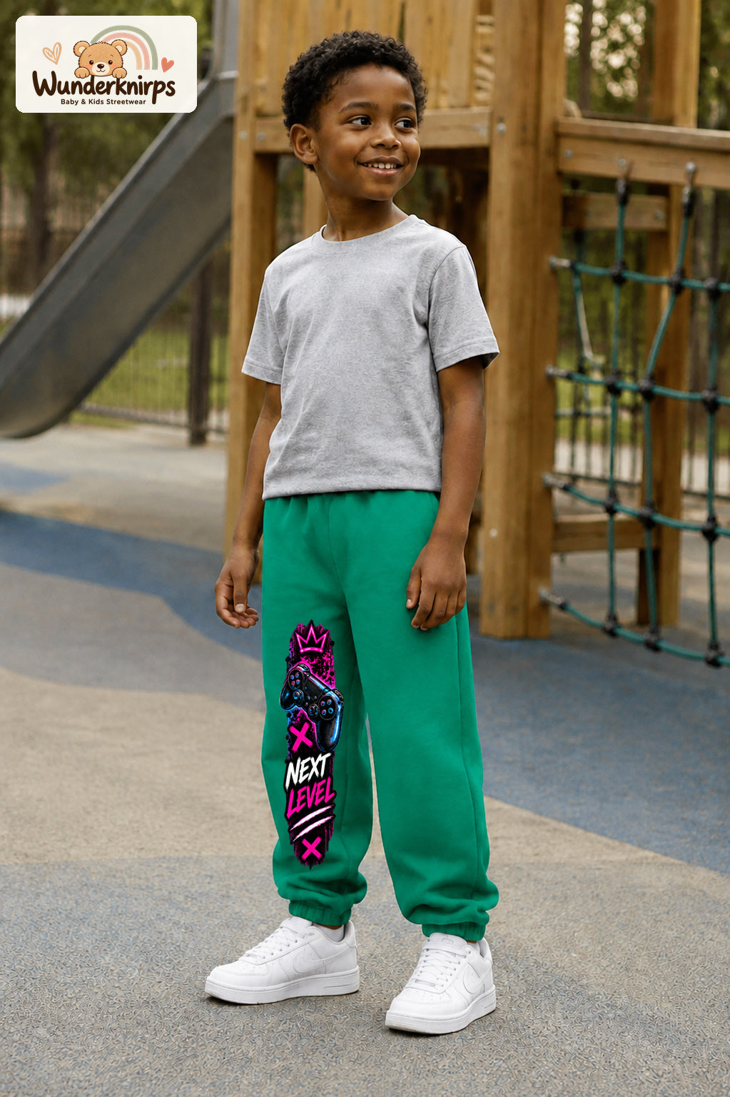 Junge trägt grüne Jogginghose mit Next Level Controller-Print, leicht seitlich auf dem Spielplatz.