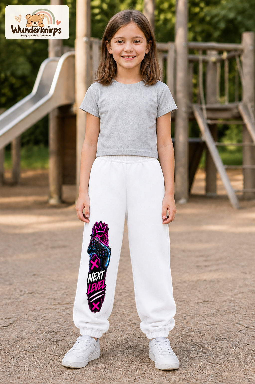 Mädchen trägt weiße Jogginghose mit Next Level Controller-Print, von vorn auf dem Spielplatz.